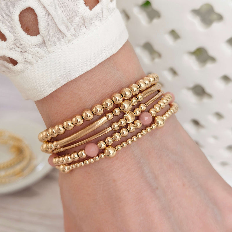 Gold-filled beaded stacking bracelets with one gold and pink opal stacking bracelet, on a wrist with a blurred neutral background