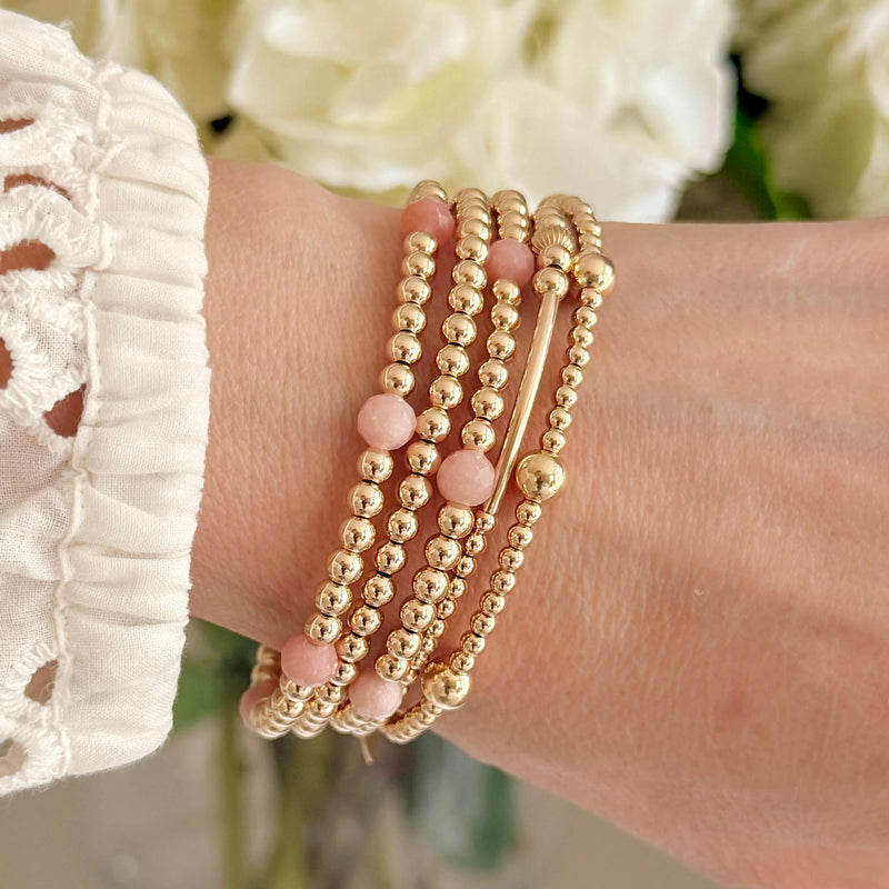 Gold-filled beaded stacking bracelets, with some featuring pastel pink opal beads, on a wrist with a blurred floral background