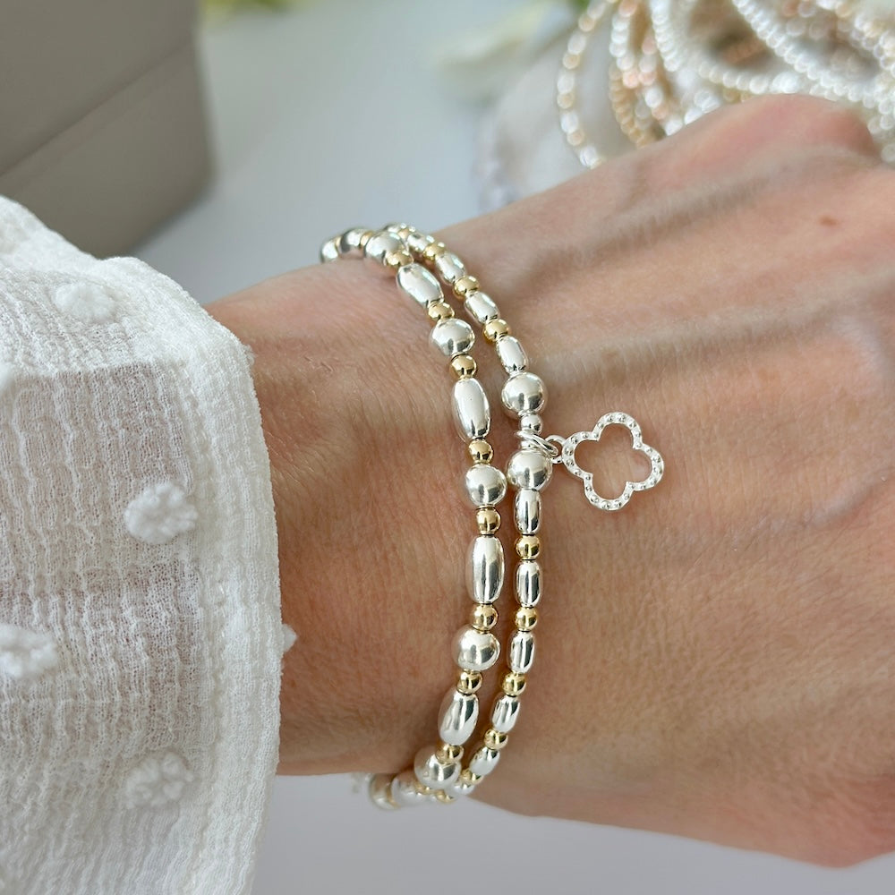 Silver and gold mixed metal, beaded bracelet duo with an open style studded four leaf clover on a wrist, with blurred jewellery in the background.