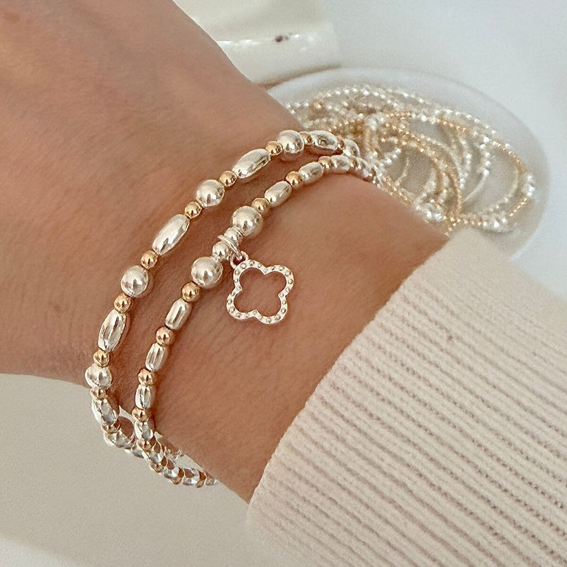 Close-up of a wrist wearing two sterling silver and gold-filled beaded stacking bracelets, one featuring a silver open clover charm, on a light background