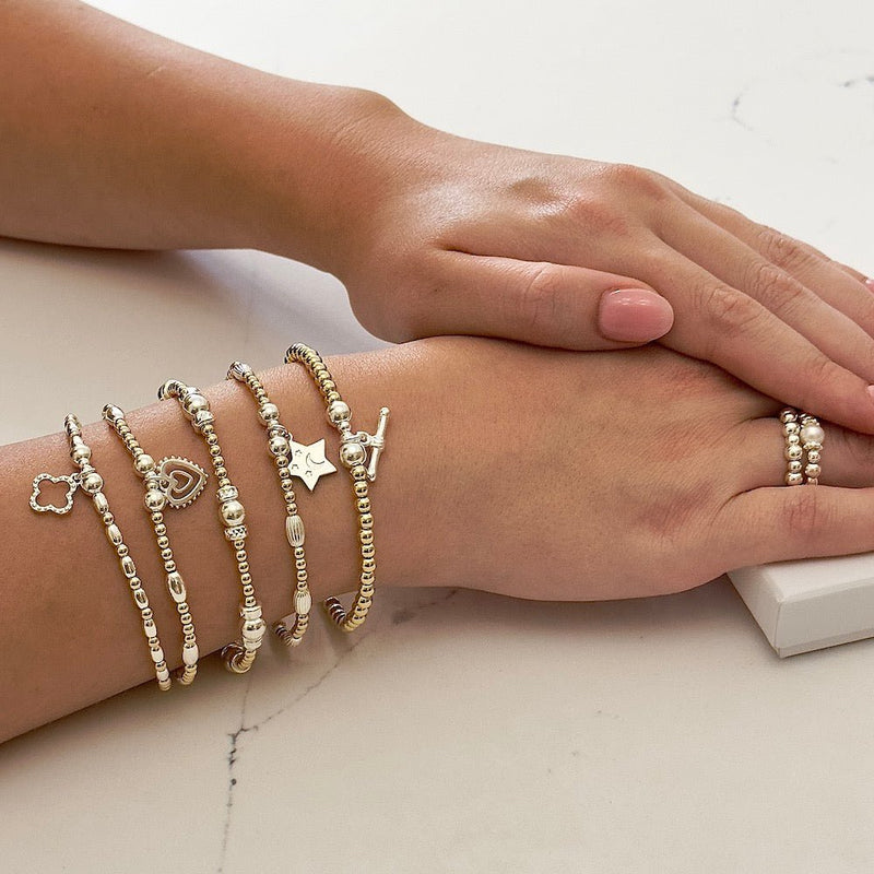 Close-up of a wrist wearing five silver and gold mixed metal beaded stacking bracelets on a light background.