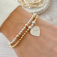 Two beaded bracelets on a wrist, one with a heart-shaped tag engraved with 'Mum'.