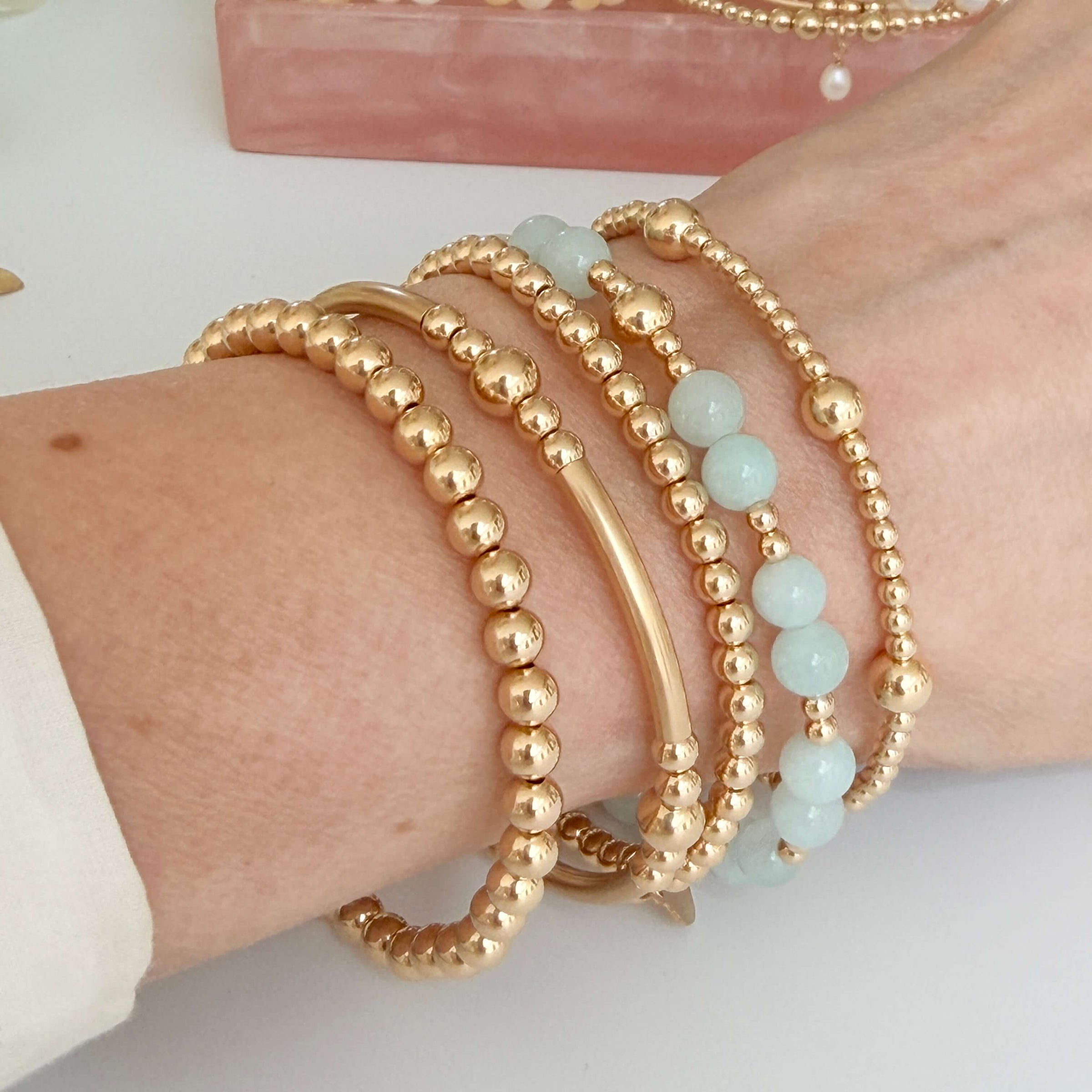 Gold beaded bracelets on a wrist with a white background, with one bracelet featuring pale blue jade beads