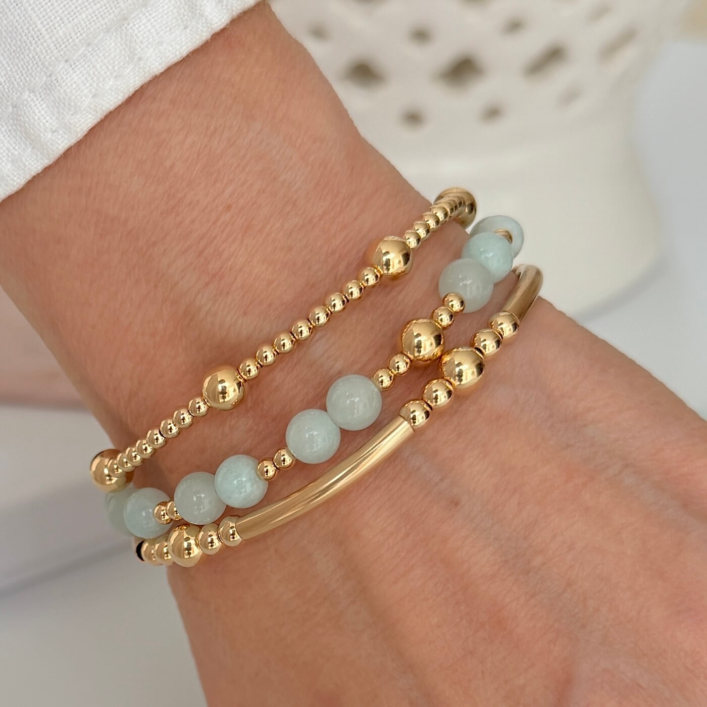 A close up of a lady's wrist wearing 3 gold stacking bracelets, one with pale blue jade beads, against a neutral background.