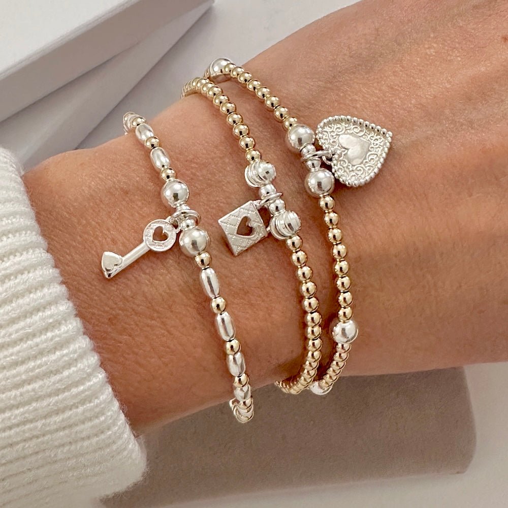 Close-up of a wrist wearing three gold and  silver mixed metal beaded bracelets with padlock, key and heart charms, on a neutral background