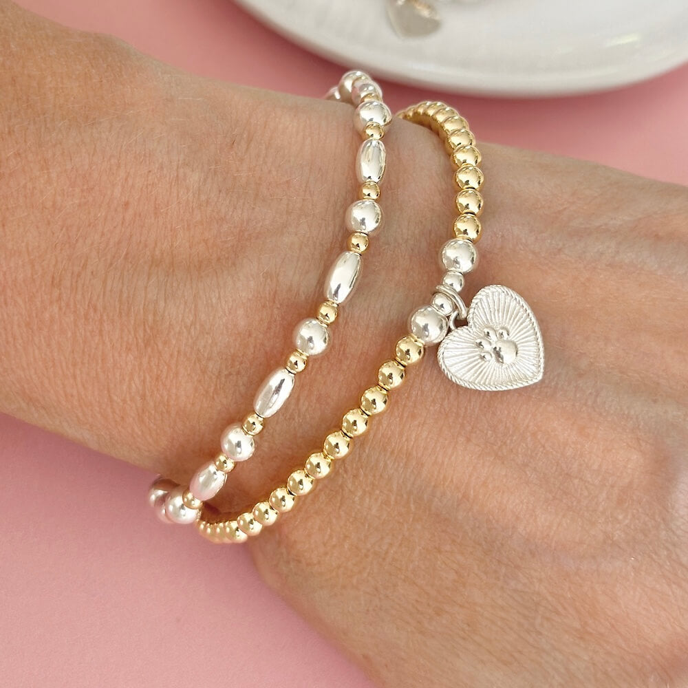 Gold and silver mixed metal beaded stacking bracelet duo with a heart charm on a lady's wrist against a pink background