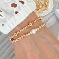 Two gold beaded bracelets, one with a pearl accent on a wrist, with a white lace top against a neutral background.