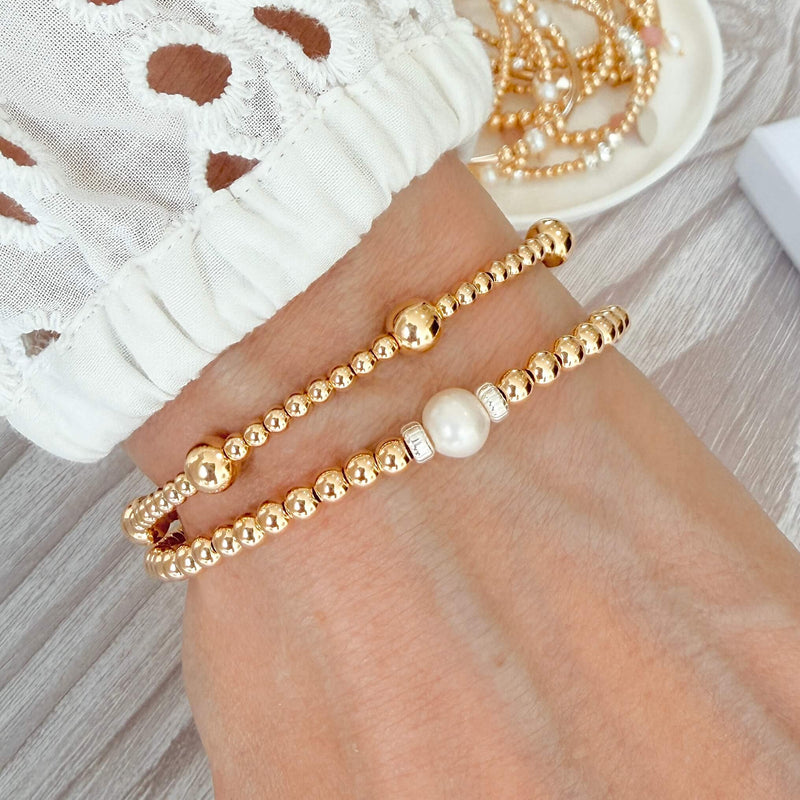 Two gold beaded bracelets, one with a pearl accent on a wrist, with a white lace top against a neutral background.