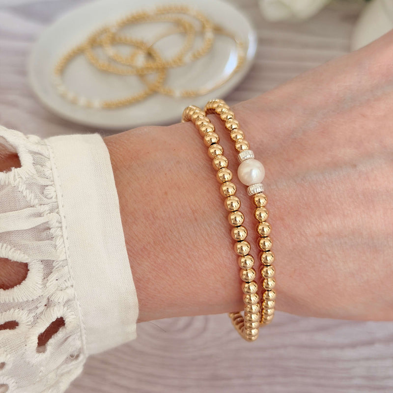 Two gold beaded bracelets, one with a pearl accent on a wrist, with a white lace top against a neutral background.