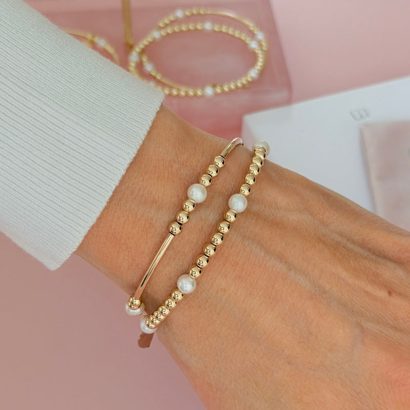 A close-up of a person's wrist wearing two gold-filled beaded bracelets with a few white freshwater pearls. A white ribbed cuff is visible, with a soft pink background and blurred jewellery packaging in the distance.