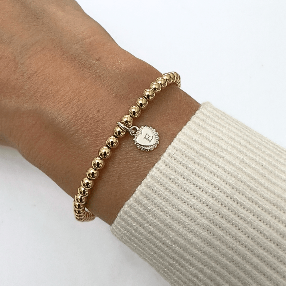 Gold beaded bracelet with a heart-shaped charm with engraved initial 'E' on a wrist against a white background