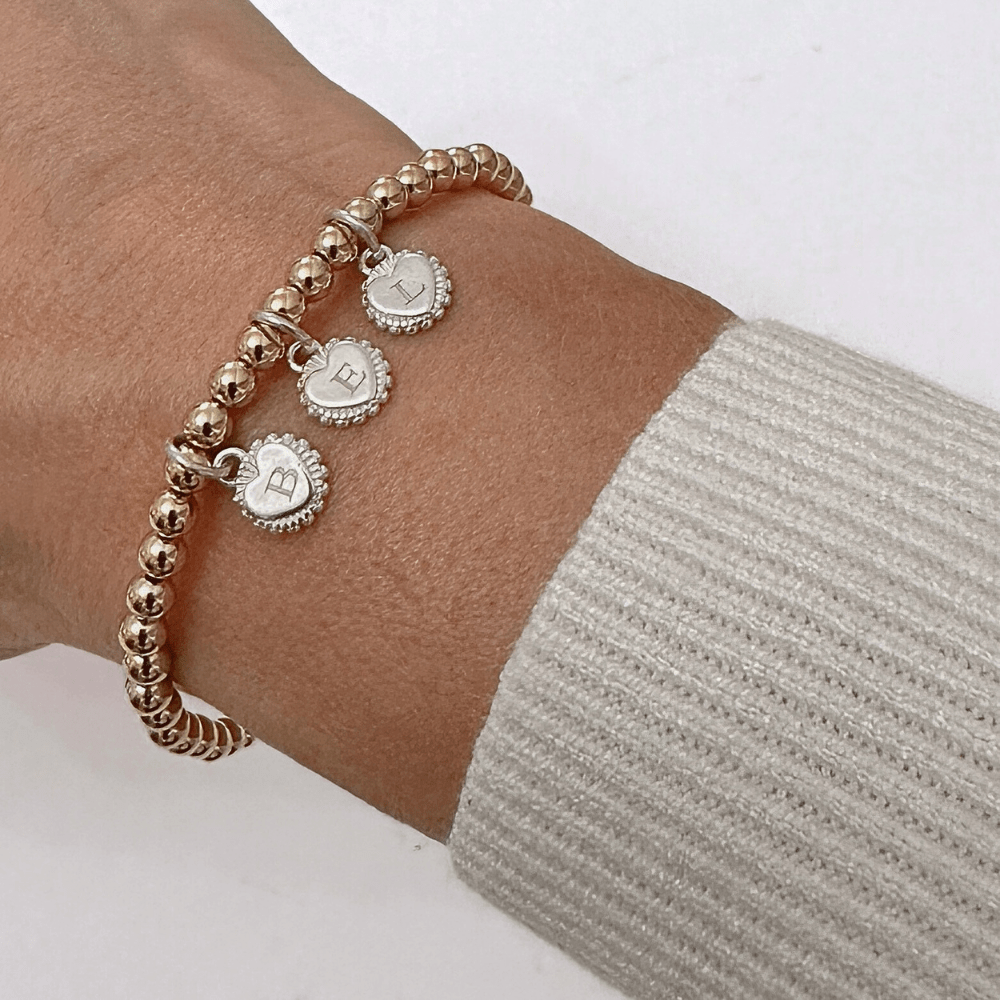 Gold bracelet with heart-shaped engraved initial disc charms on a wrist against a white background