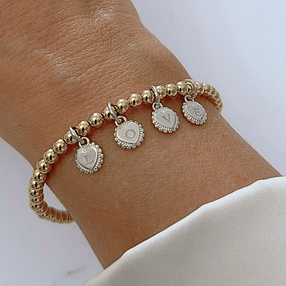 Gold beaded bracelet with four heart-shaped charms with initials engraved on them, on a wrist against a white background