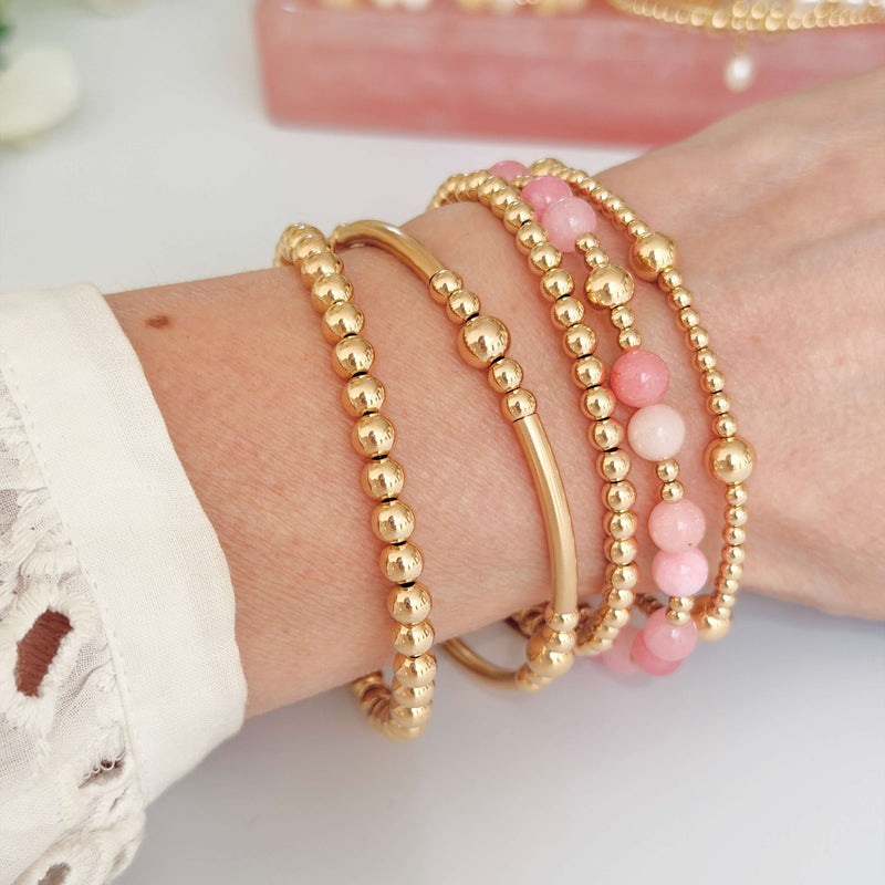 Gold beaded bracelets on a wrist, one with pale pink beads for summer, against a blurred background
