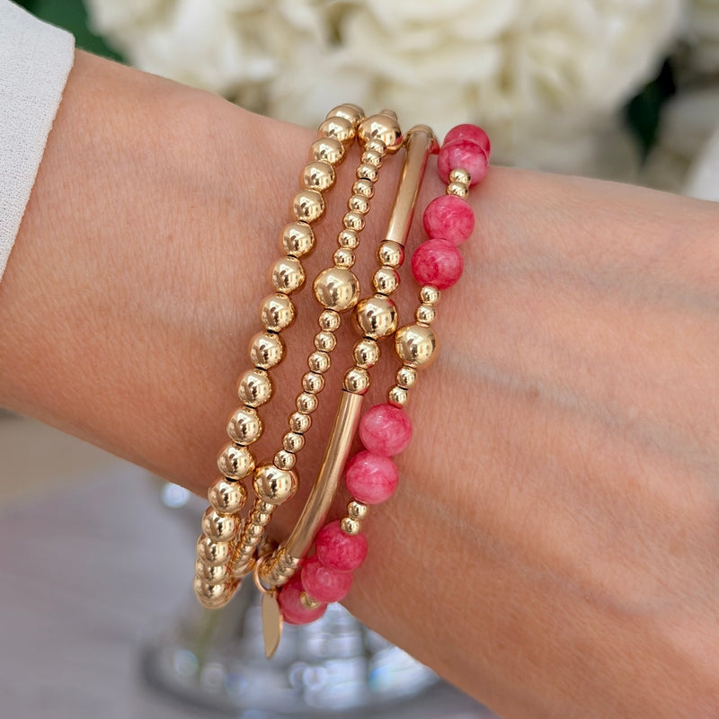 A close-up of a person's wrist wearing a stack of four beaded bracelets: three gold beaded bracelets and one beaded bracelet with raspberry pink stone beads mixed with gold beads, against a light neutral background.