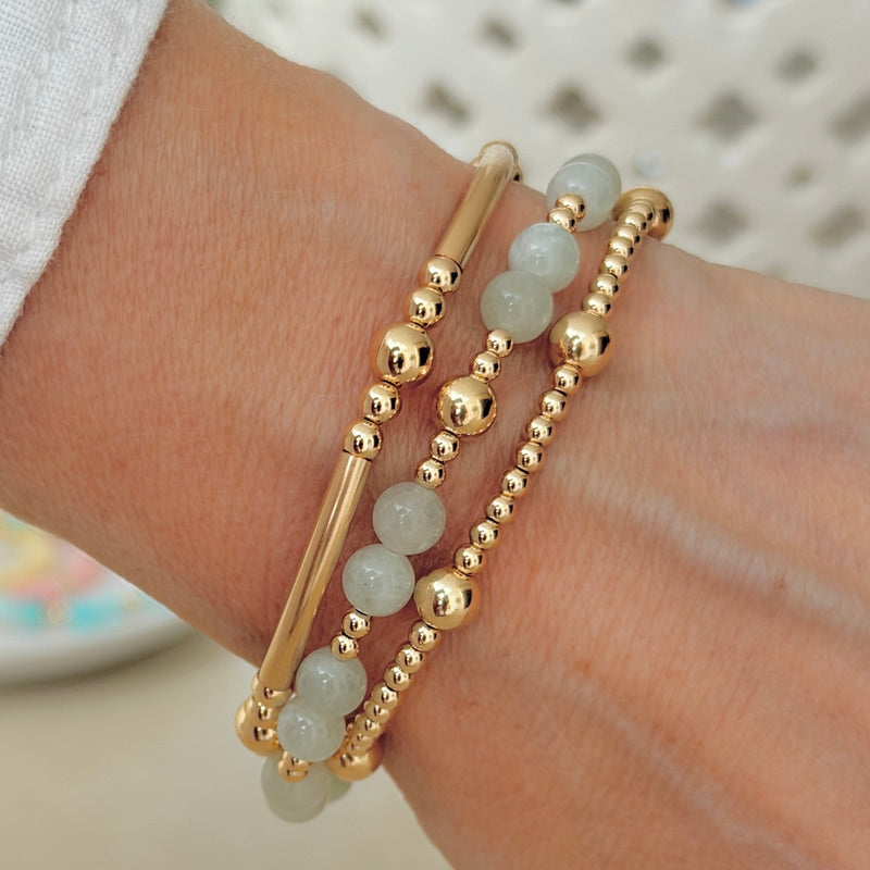 A wrist wearing two gold beaded bracelets and a third gold and pale green beaded stacking bracelet, against a blurred background.