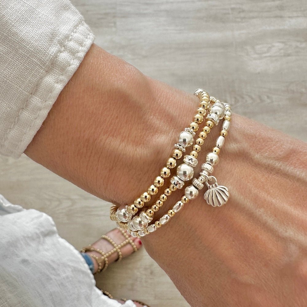Close-up of a wrist wearing multiple silver and gold beaded bracelets with a siolver shell charm.