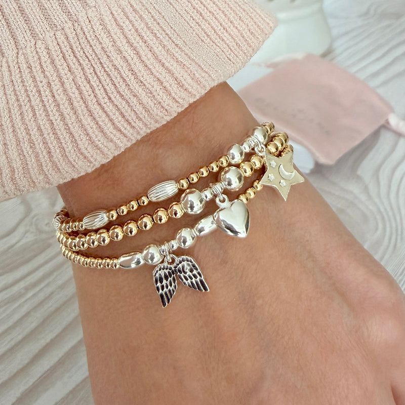 A close-up of a person's wrist wearing three sterling silver and gold-filled mixed metal beaded bracelets, featuring an angel wings charm, a puffed heart charm and a star charm, styled with a pink knitted jumper.