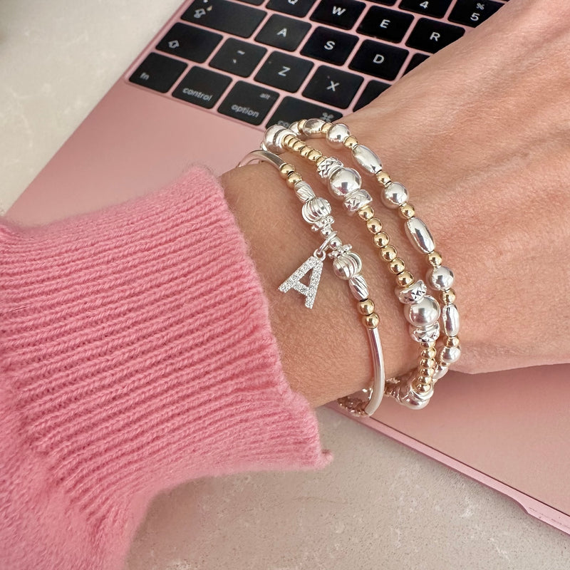 A lady's wrist wearing 3 gold and silver beaded stacking bracelets with a sparkly initial A charm and pink sleeve