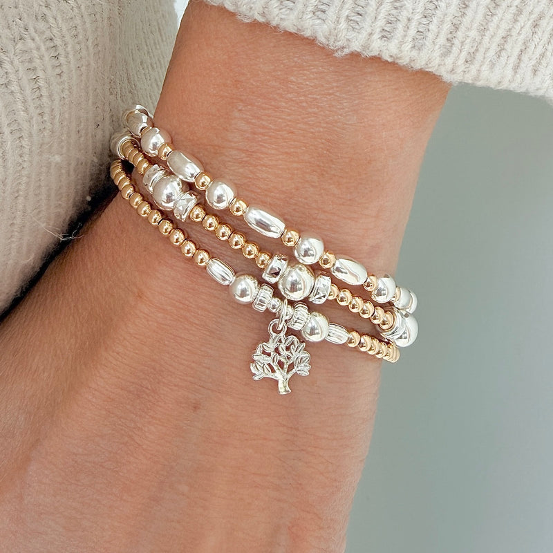 A close up of a person's wrist wearing 3 gold and silver beaded bracelets, with one featuring a small silver tree charm.