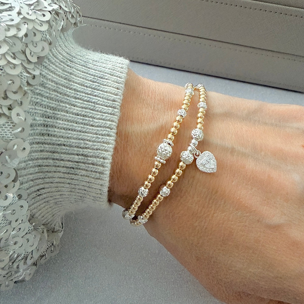 Two gold and silver mixed-metal beaded stacking bracelets, one with a heart charm on a wrist, against a neutral background
