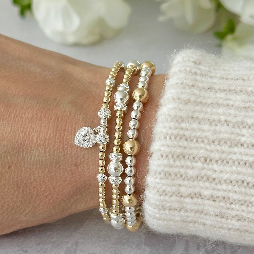 Gold and silver mixed metal beaded bracelets on a wrist, one bracelet featuring a cubic zirconia heart charm, against a light background.