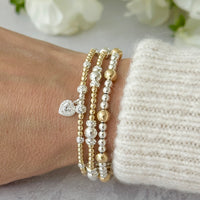 Gold and silver mixed metal beaded bracelets on a wrist, one bracelet featuring a cubic zirconia heart charm, against a light background.