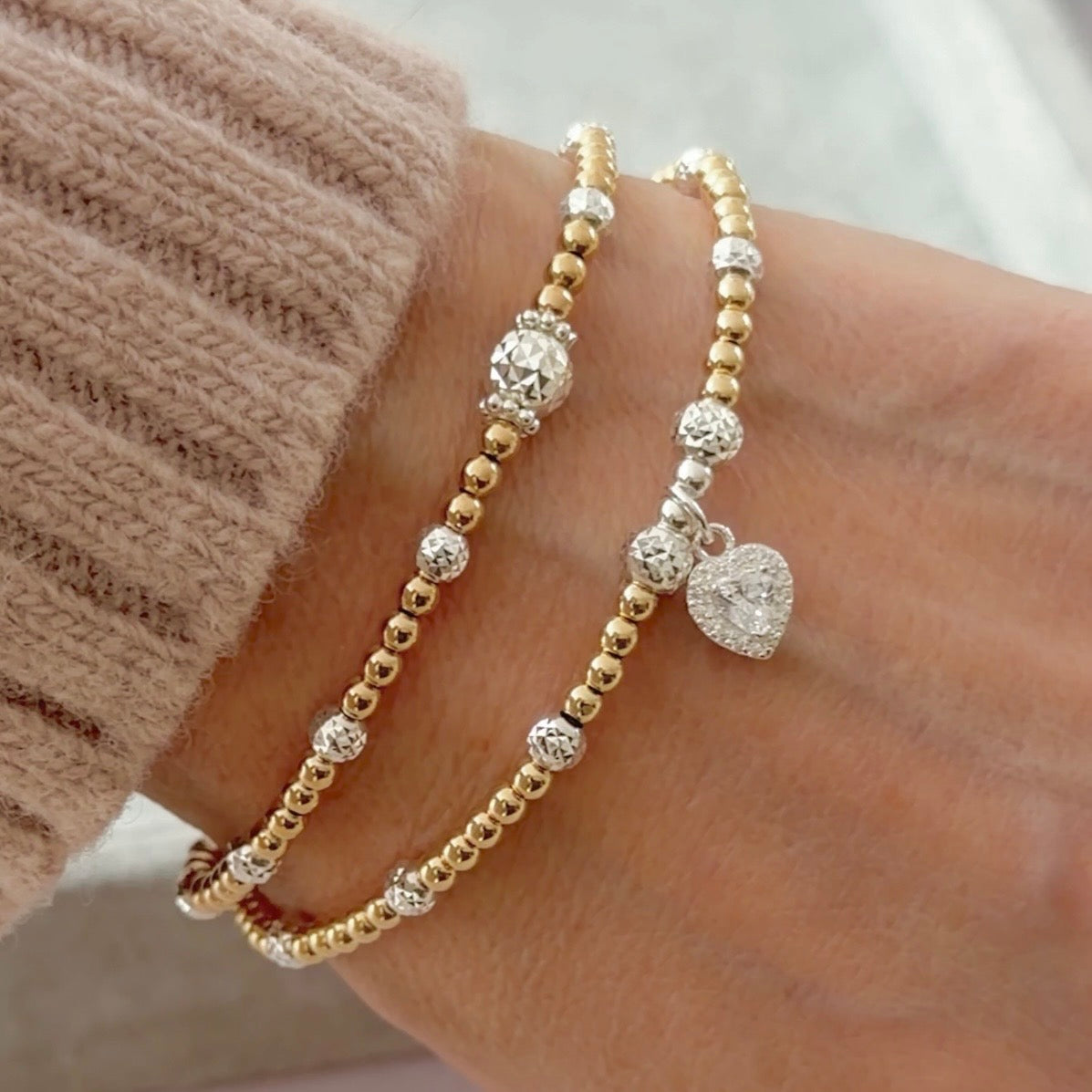 A close up of a wrist wearing two gold and silver mixed metal beaded stacking bracelets, one featuring a sparkly heart charm against a neutral background.