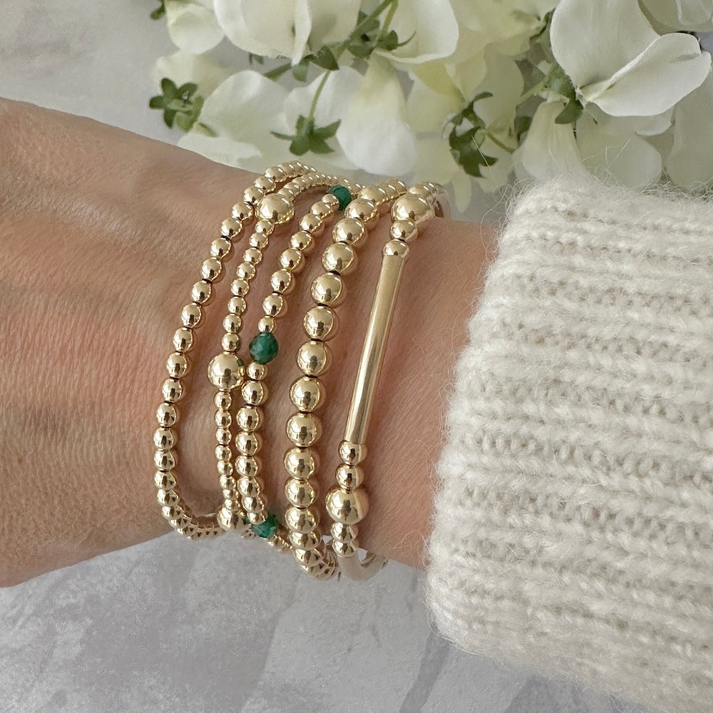 Gold beaded bracelets on a wrist, one bracelet featuring green onyx beads, against a light background