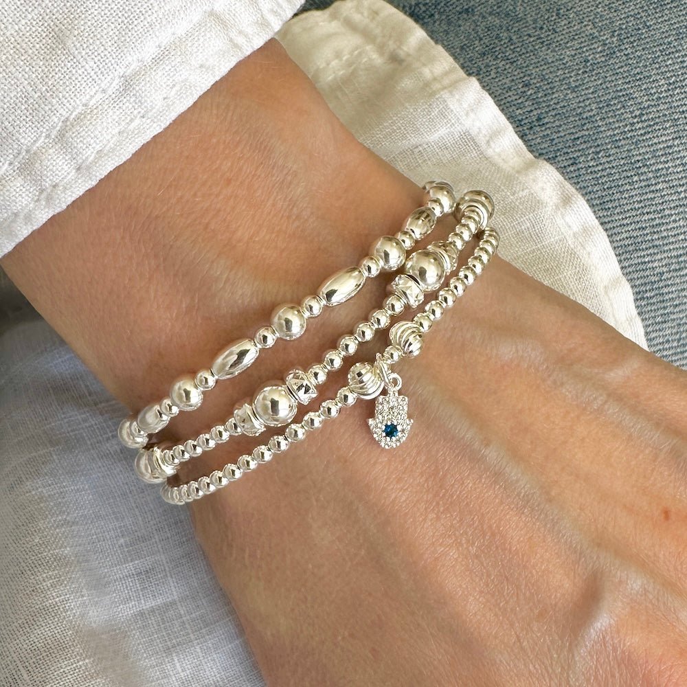 A close up of a lady's wrist wearing a trio of sterling silver beaded bracelets, one with a sparkly and blue gemstone hand of protection charm, against a neutral background