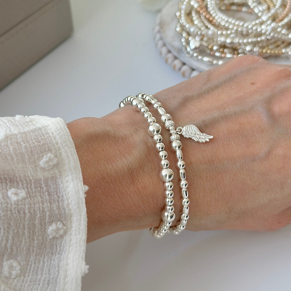 Silver bracelet duo with angel wing charm on a wrist, with a blurred image of stacking bracelets in the background.