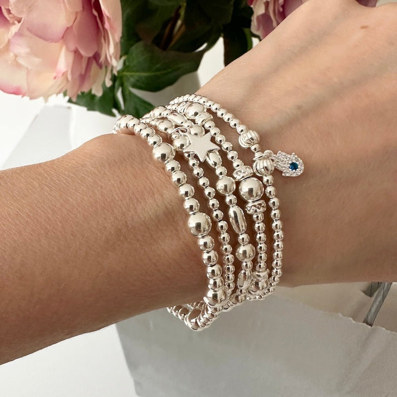 Silver beaded bracelets on a wrist with a small sparkly hand charm, against a floral background