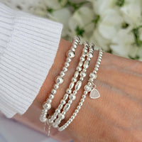 Silver beaded bracelets on a wrist, one featuring the June birthstone charm, with a blurred floral background