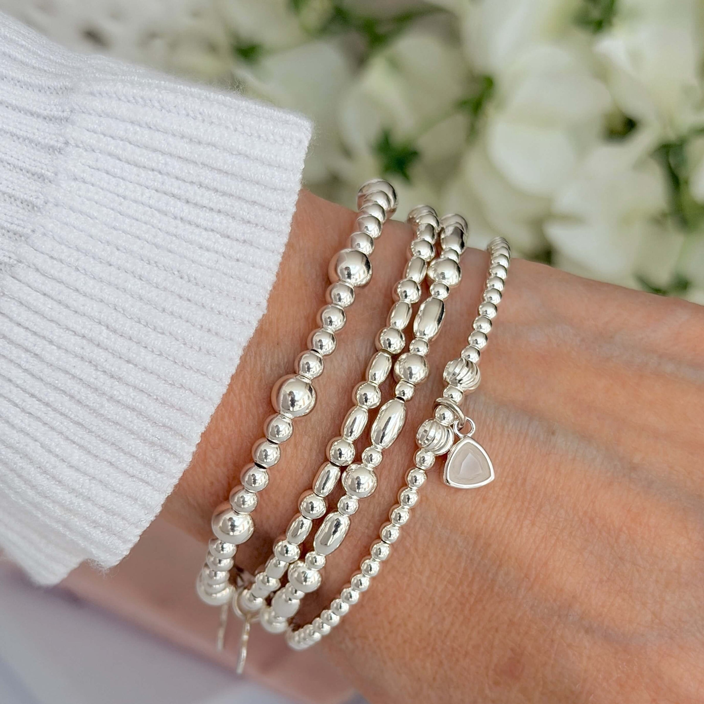 Silver beaded bracelets on a wrist, one featuring the June birthstone charm, with a blurred floral background