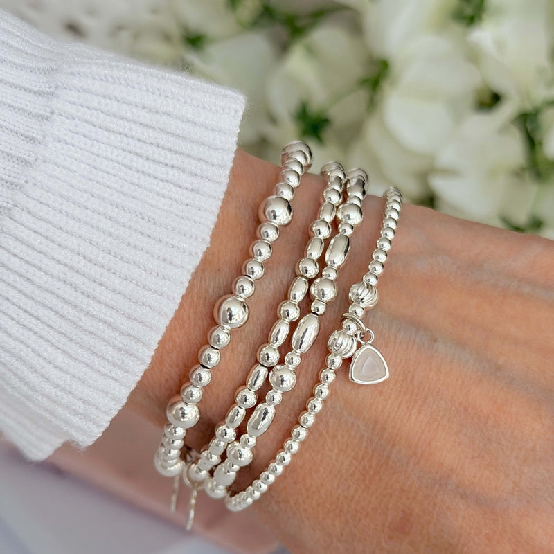 Silver beaded bracelets on a wrist, one featuring the June birthstone charm, with a blurred floral background