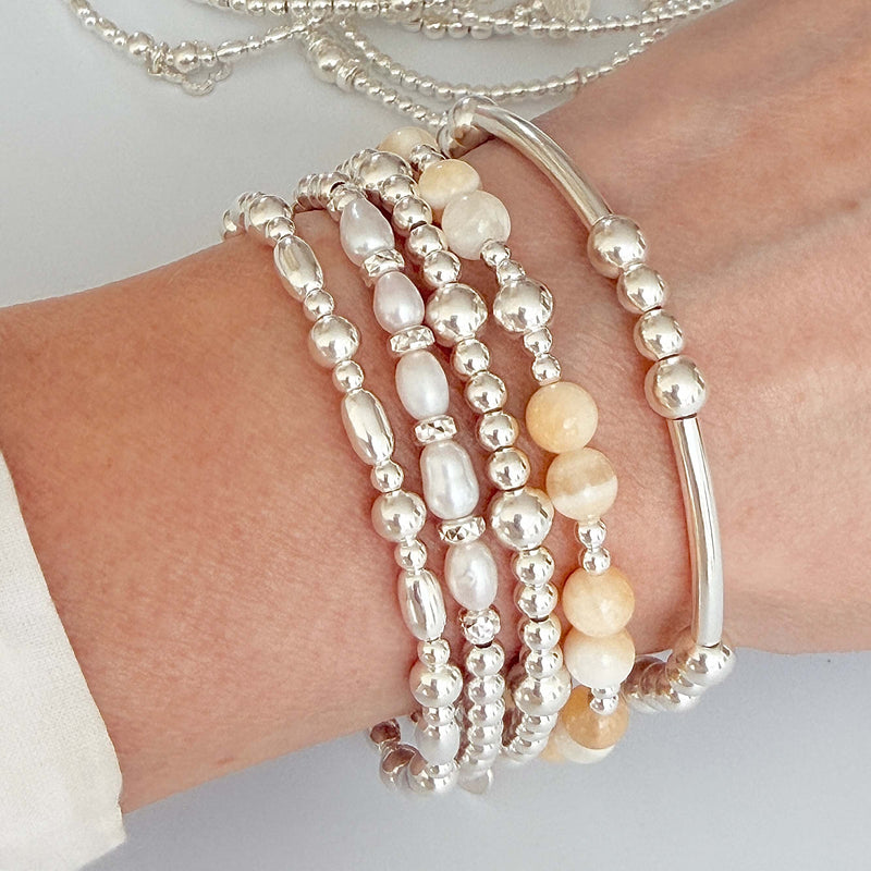 Close-up of a wrist wearing multiple sterling silver bracelets with a pale yellow beaded bracelet on a light background