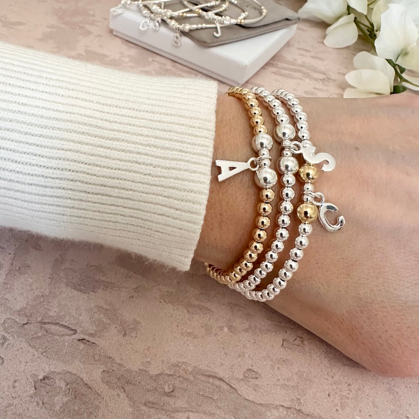 A close up of a lady's wrist featuring 3 Bella Jane beaded stacking bracelets in a mix of sterling silver and gold-filled beads, with different sterling silver initial charms, against a neutral background.