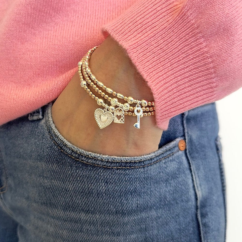 Gold and silver mixed-metal stacking bracelets with heart and key charms on a person wearing a pink sweater and blue jeans.