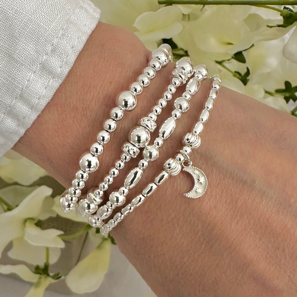 Silver beaded bracelets on a wrist with a floral background, one bracelet featuring a sterling silver and ivory enamel moon charm, with tiny stars