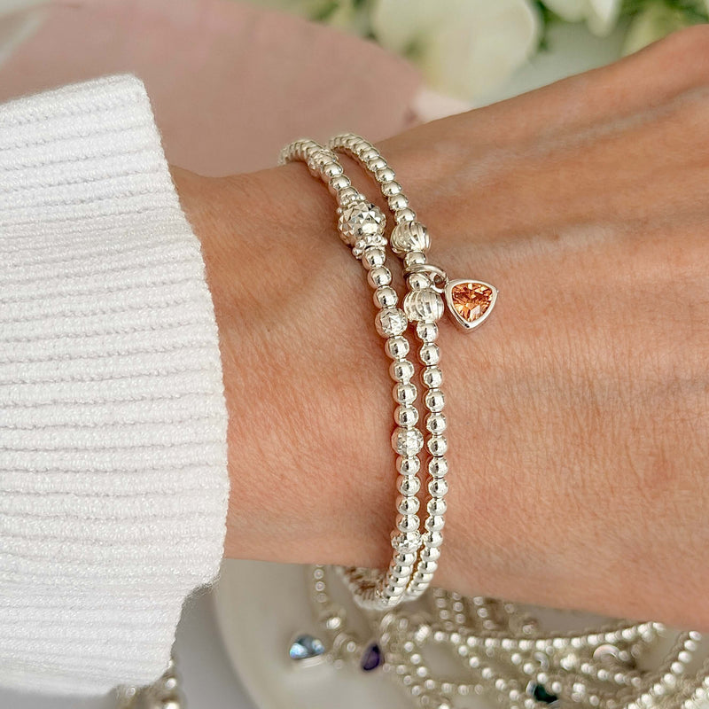 A ladies wrist wearing two silver beaded bracelets, one featuring a November birthstone charm, on a wrist against a blurred floral background