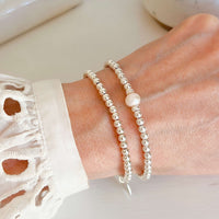 Two beaded silver stacking bracelets, one featuring a pearl in the centre, on a lady's wrist with a blurred background