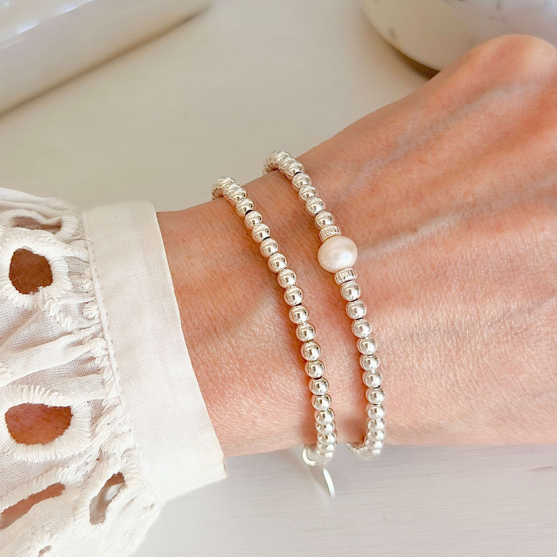 Two beaded silver stacking bracelets, one featuring a pearl in the centre, on a lady's wrist with a blurred background