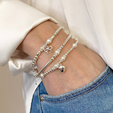 Close-up of a hand wearing multiple silver and freshwater pearl bracelets with heart and bow charms with an ivory silk shirt sleeve with hand in jeans pocket.