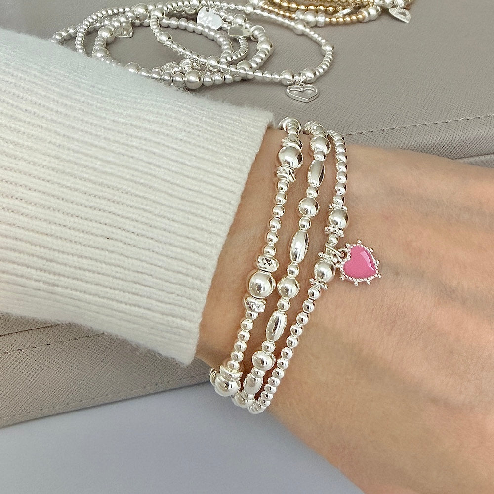 Silver beaded bracelets on a wrist with one featuring a pink heart charm, against a neutral background.