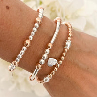 Three mixed-metal sterling silver and rose gold stacking bracelets on a wrist with a blurred floral background