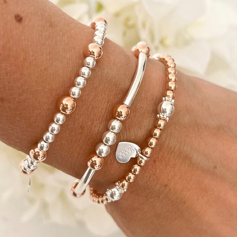 Three mixed-metal sterling silver and rose gold stacking bracelets on a wrist with a blurred floral background