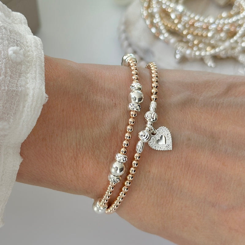 Close-up of a wrist wearing two beaded rose gold and silver mixed metal bracelets, with heart charm on a neutral background