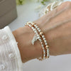 Silver and rose gold mixed metal bracelet duo with angel wing charm on a wrist, with a blurred image of stacking bracelets in the background.