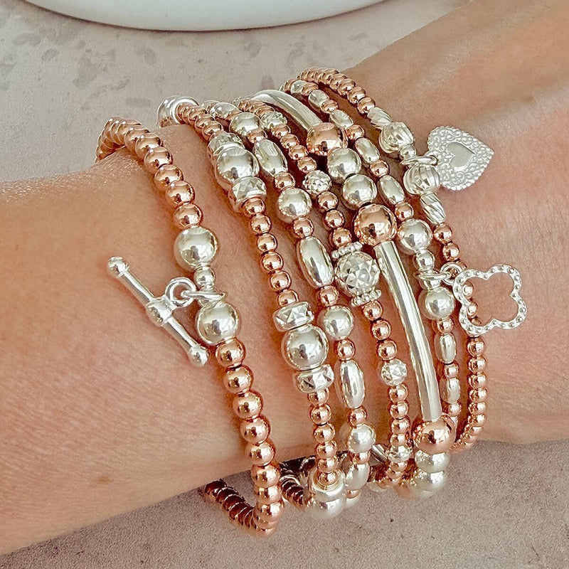 A close-up of a person's wrist wearing a stack of beaded bracelets in rose gold and silver tones, including round beads, polished metal beads and charms.