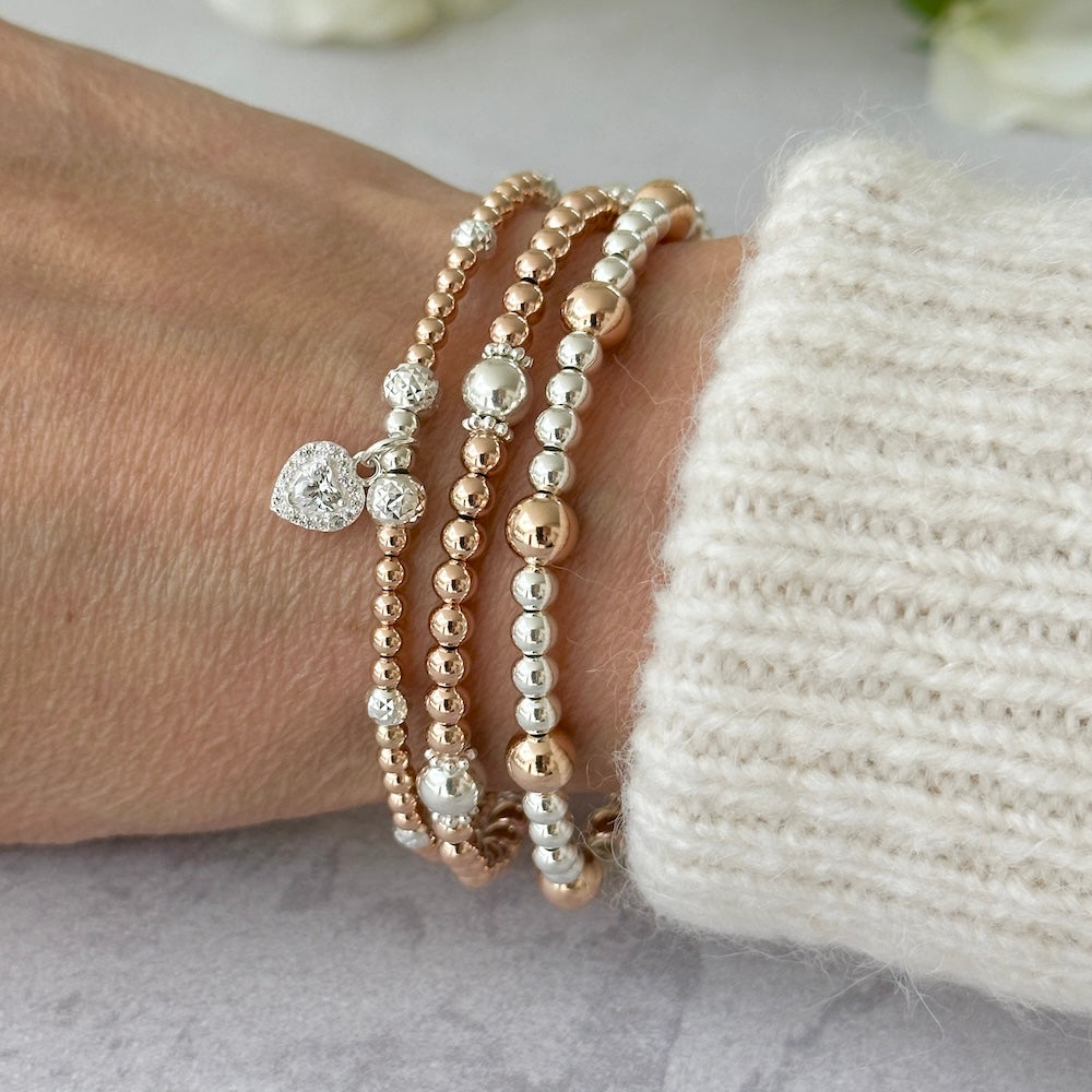 Rose gold and silver mixed metal beaded bracelets on a wrist, one bracelet featuring a cubic zirconia heart charm, against a light background.